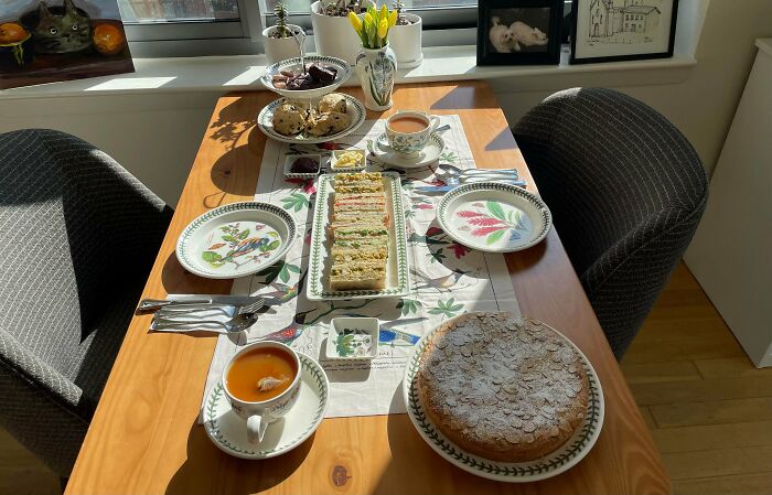 Cozy cottage core aesthetics with floral teacups, homemade cake, sandwiches, scones, and fresh flowers on a wooden table.