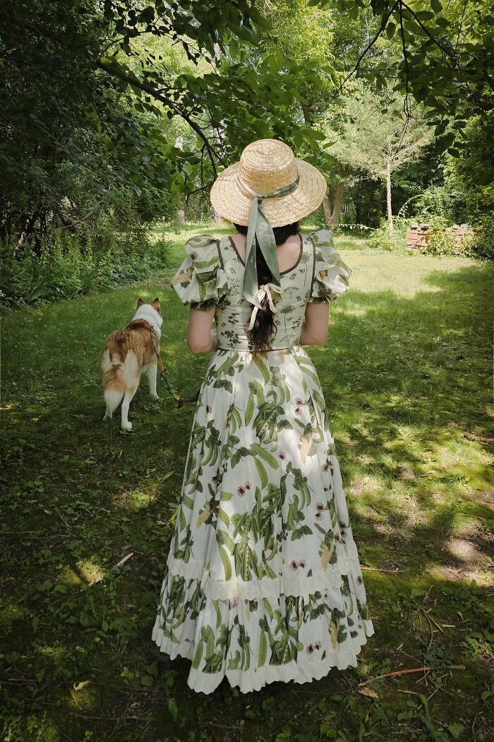 Woman in a floral cottage core dress with a straw hat walking a dog in a lush green garden setting.