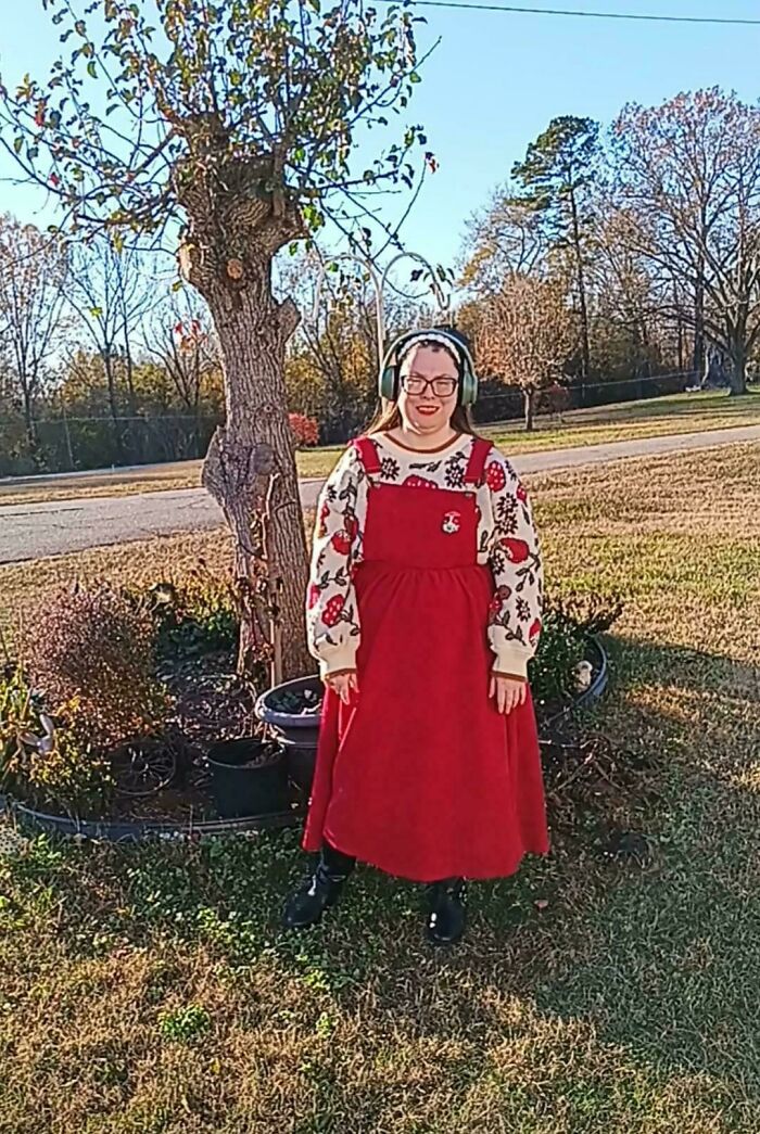 Woman wearing a red pinafore dress and floral sweater standing outdoors in a sunny setting showcasing cottage core aesthetics.