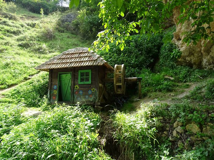 Small wooden cottage with floral decorations and water wheel surrounded by lush greenery and natural landscape in cottage core aesthetics.