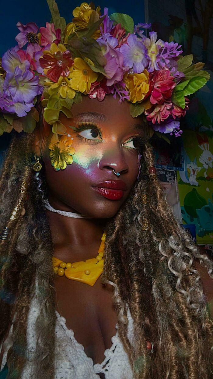 Young woman with colorful flower crown and glowing makeup, embodying magical cottage core aesthetics with natural braided hair.