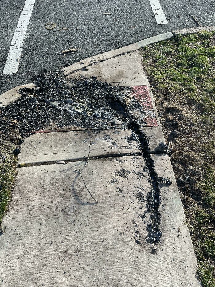 Burnt debris on a sidewalk corner next to a parking lot and grass, showing unexpected urban damage and cleanup efforts.