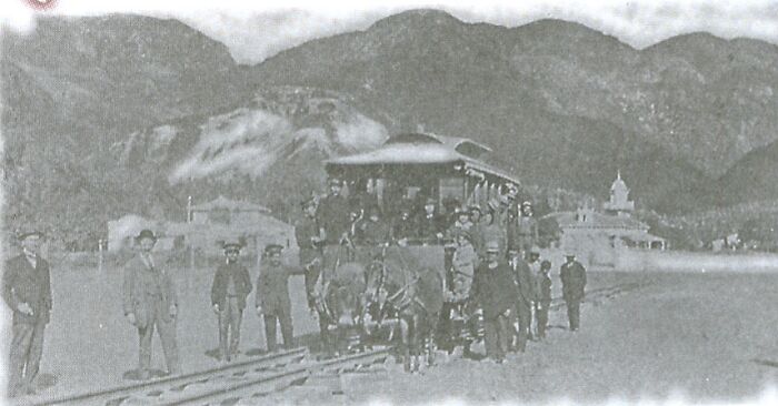Historic black and white photo showing an early train with people on and around it in a world capital before modern times.
