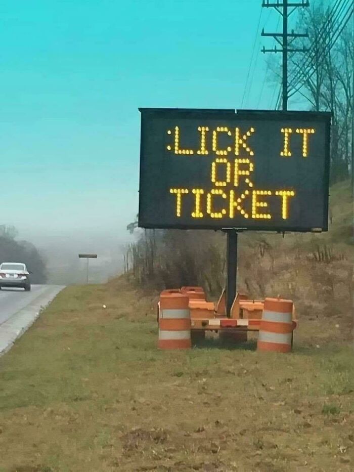 Portable LED sign on roadside displaying the message lick it or ticket, surrounded by orange traffic barrels.