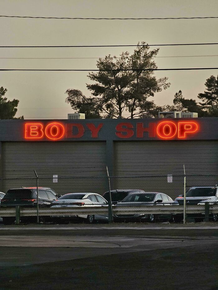 Neon LED sign displaying body shop on a commercial building with parked cars and trees in the background.