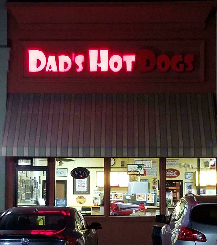 LED sign glowing in red for Dad's Hot Dogs above a restaurant entrance at night with parked cars visible.