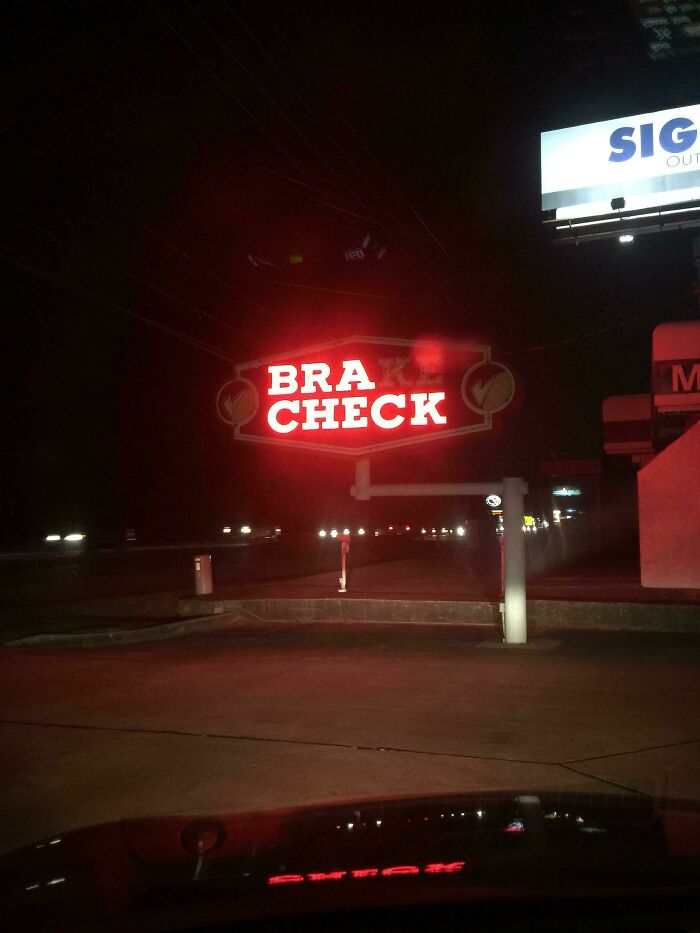 Red LED sign glowing at night displaying the words bra check on a roadside near a gas station.