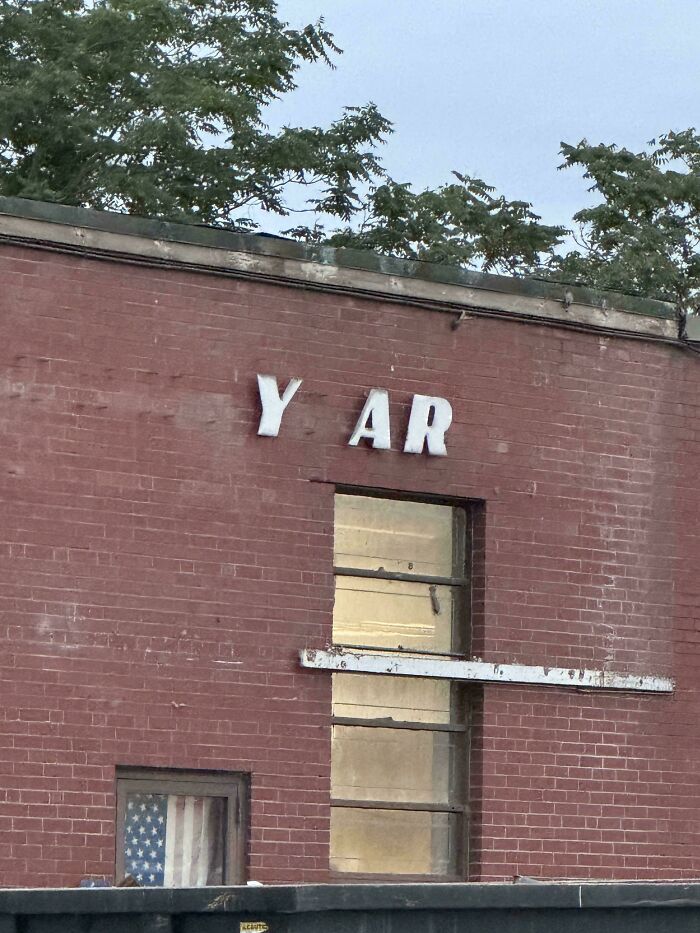 Partial LED sign on a brick wall missing letters, showing only Y AR with trees and a window in the background.