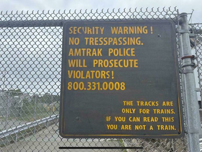 Security warning sign on chain link fence with a humorous message about trespassing on train tracks.