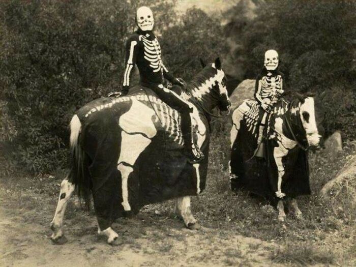 Two people wearing vintage skeleton Halloween costumes while riding horses dressed in matching skeleton designs outdoors.