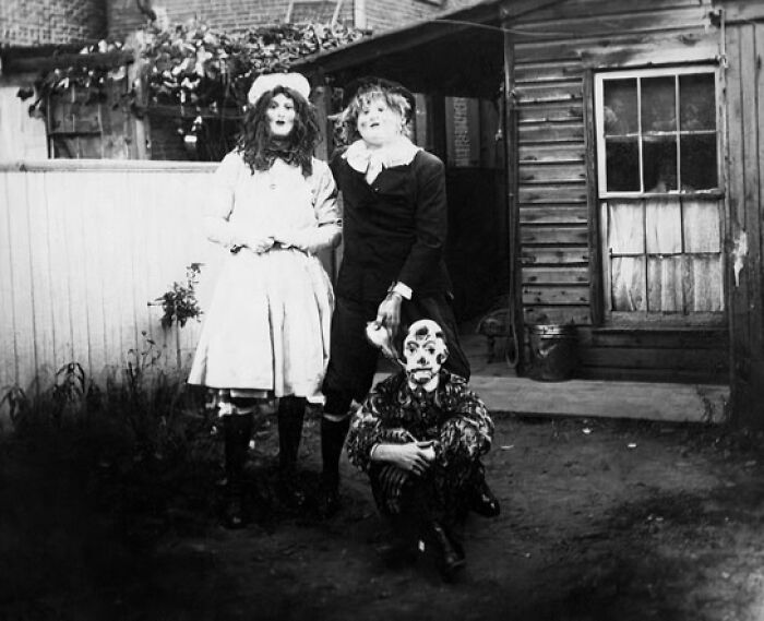 Three people wearing vintage Halloween costumes, including a clown mask, posing outdoors in front of a wooden house.