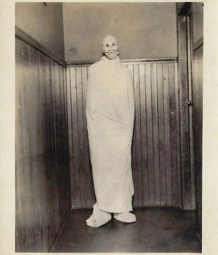 Vintage Halloween costume showing a person wrapped in white cloth and wearing a creepy skull mask in a wooden-paneled room.