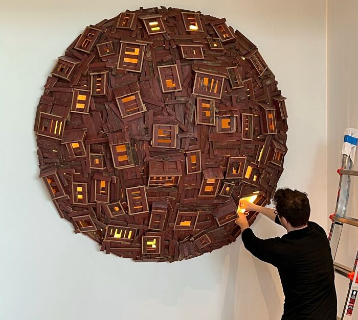 Artist adjusting a surreal collage artwork of walking and collapsing buildings with illuminated windows on a white wall.