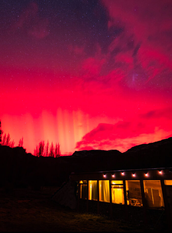 Vibrant red aurora lighting the night sky above a warmly lit cabin in a remote landscape, interesting pics nature scene.