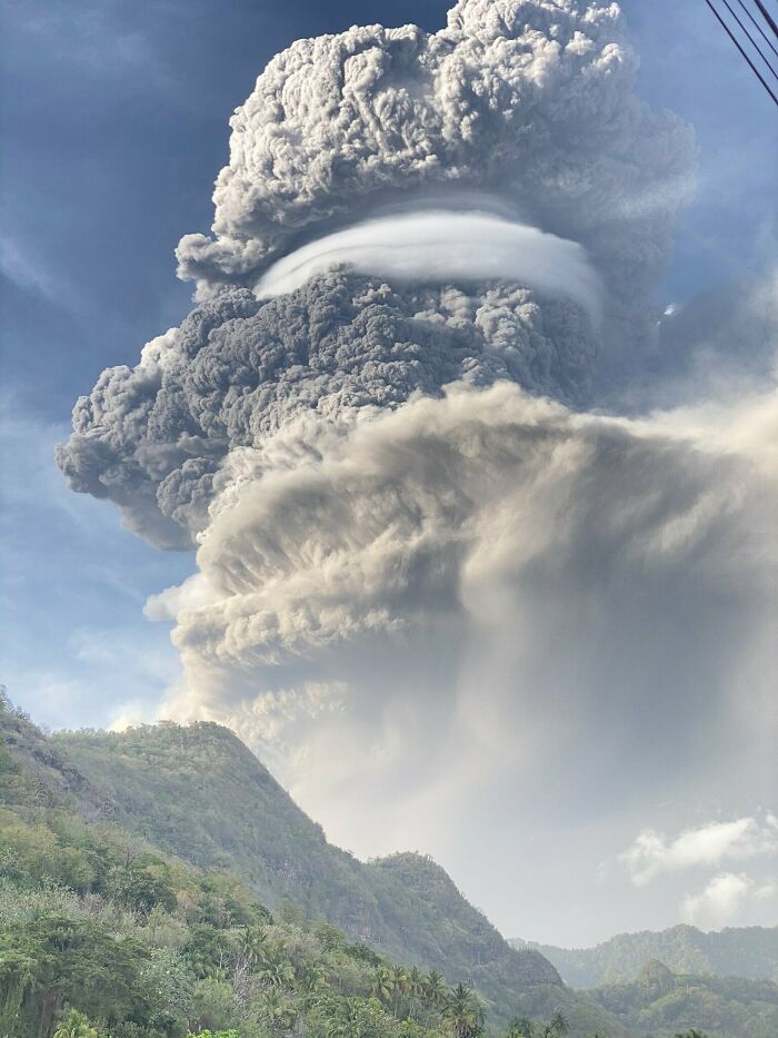 Massive volcanic eruption with towering ash clouds over lush green mountains in an interesting pic answering questions.