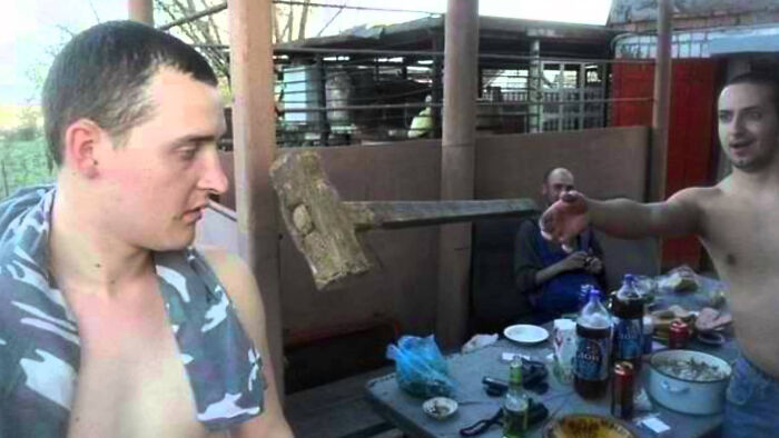 Two shirtless men at a table with drinks, one unaware of a hammer flying toward his head before disaster struck.