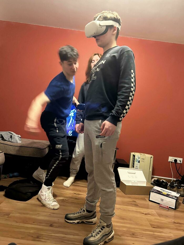 Three friends in a room with one wearing VR goggles while another is about to accidentally collide, capturing a hilarious disaster moment.