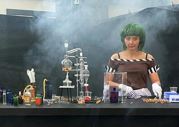 Woman with green wig conducting a chemistry experiment with colorful liquids and glassware in a smoky setting, pop culture moments.