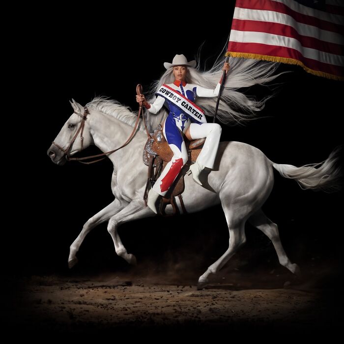 Person in a Cowboy Carter outfit riding a white horse holding an American flag, showcasing pop culture moments 2024.