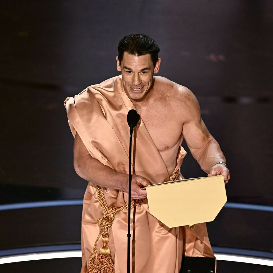 Actor in a toga-style outfit speaking at a microphone holding an envelope during a 2024 pop culture moments event.