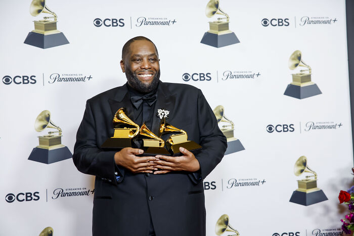 Man in black suit holding multiple Grammy awards, celebrating a surprising pop culture moment in 2024.