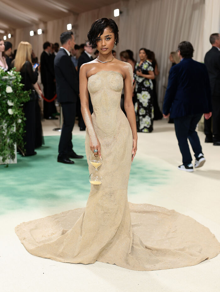 Woman in a sand-colored gown holding an hourglass at a formal event, highlighting unexpected 2024 pop culture moments.