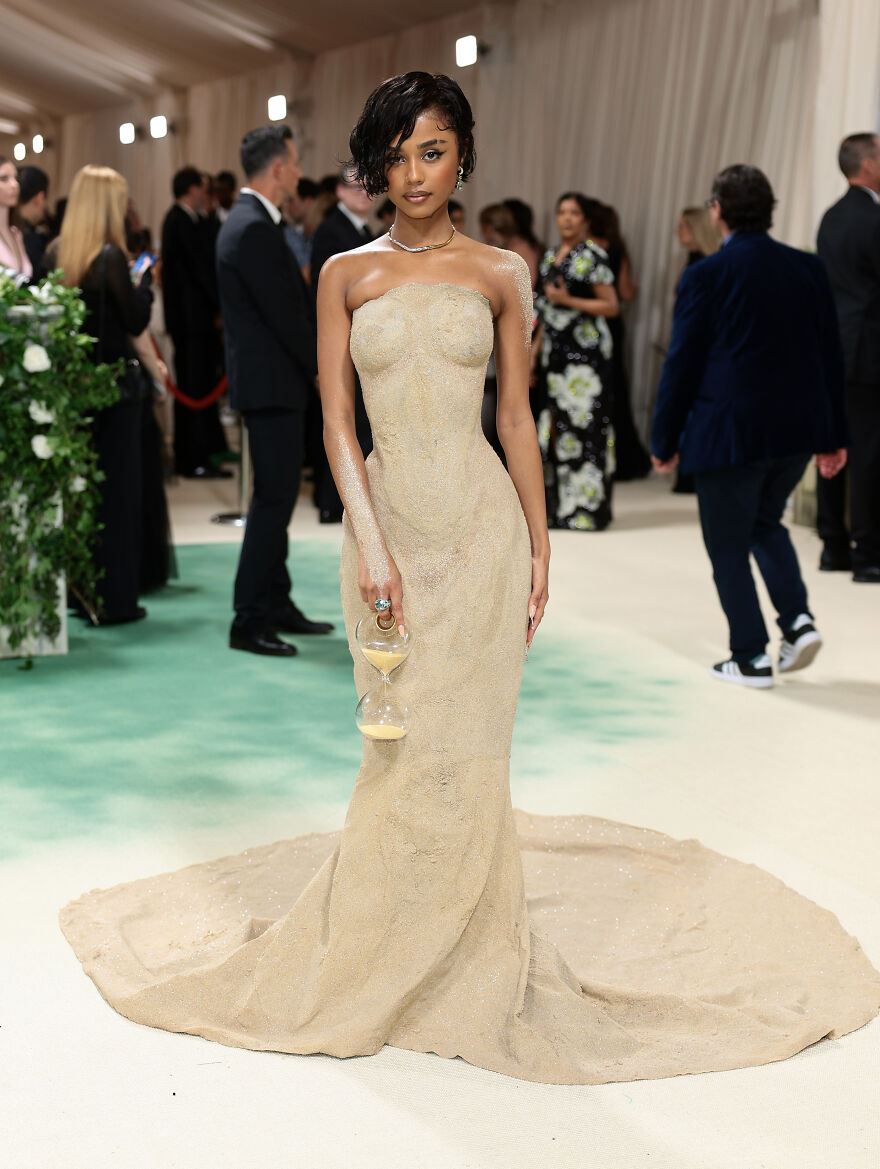 Woman in an elegant beige gown holding an hourglass at a glamorous event, highlighting 2024 pop culture moments that shocked fans.