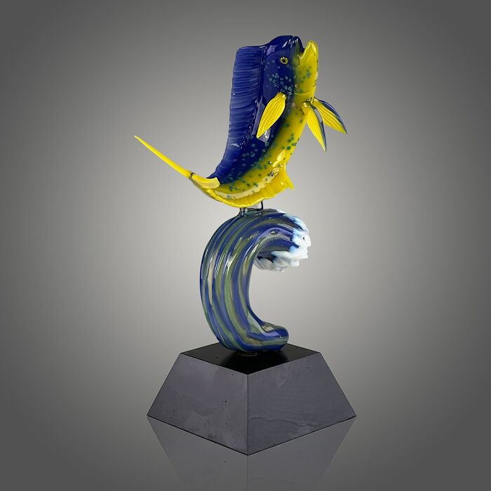 Glass sculpture of a vibrant yellow and blue fish jumping over a stylized ocean wave on a black pedestal.