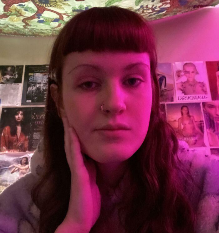 Young woman with uneven blunt bangs and long hair, lit by pink light, posing indoors with posters in background showing hair accidents.