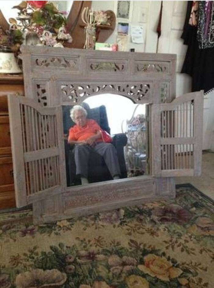 Ornate wooden mirror with shutters reflecting an elderly person sitting on a chair in a cozy, decorated room.