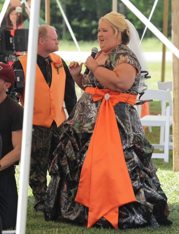Bride wearing a camouflage wedding dress with bright orange sash and groom in matching vest at outdoor wedding ceremony.