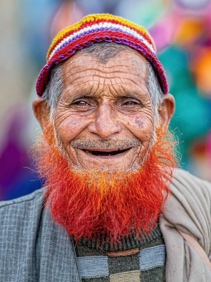 Elderly man with vibrant orange beard and colorful cap smiling, capturing the beauty of everyday life in India by street photographer.