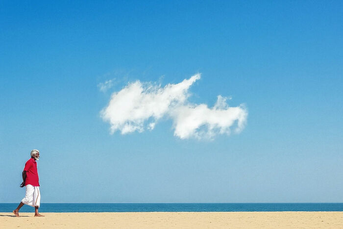 Elderly man walking on a serene beach under a blue sky with clouds, capturing everyday life beauty in India.
