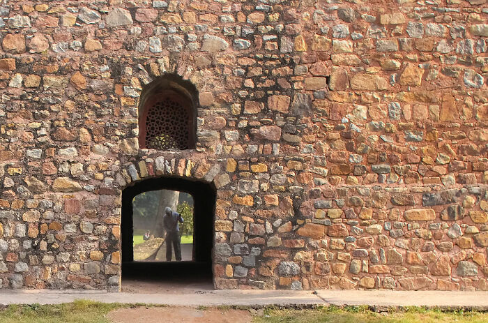 Man sweeping ground through arched doorway of historic stone wall, capturing beauty of everyday life in India.