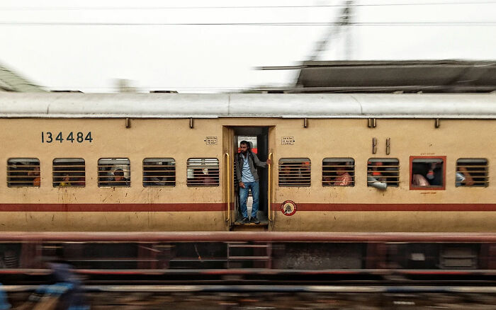 Man standing in doorway of moving train with passengers inside, capturing the beauty of everyday life in India street photography.