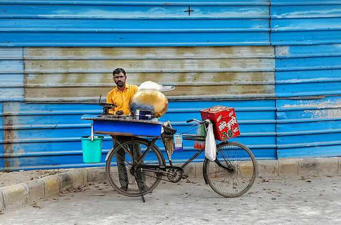 Street photographer captures beauty of everyday life in India with a man selling snacks from a bicycle cart against blue wall.