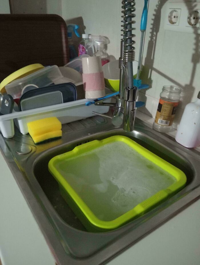 Kitchen sink with soapy water in a yellow basin and various cleaning supplies on a drying rack nearby in an interesting group setting.