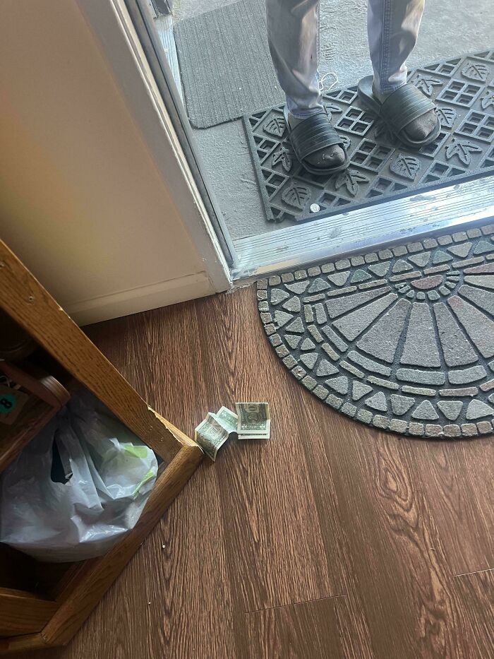 Person wearing sandals standing outside a door with crumpled dollar bills trapped under the door frame on a wooden floor.