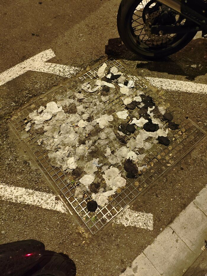 Grate on asphalt covered with black and white debris, surrounded by parking lines and partial motorcycle view at night.