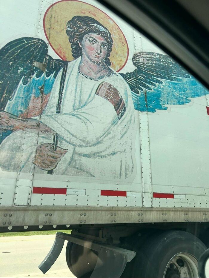 Mural of an angel with blue wings and a halo painted on the side of a semi-truck in an unusual roadside scene.