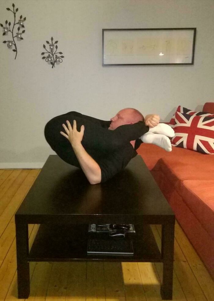 Man demonstrating an unexplained flexible pose on a coffee table in a living room with a Union Jack pillow.