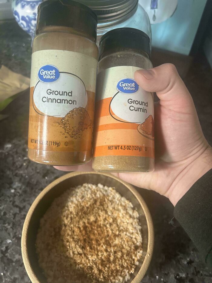 Hand holding ground cinnamon and ground cumin spice bottles over a bowl of oatmeal, showing a messy kitchen scene.