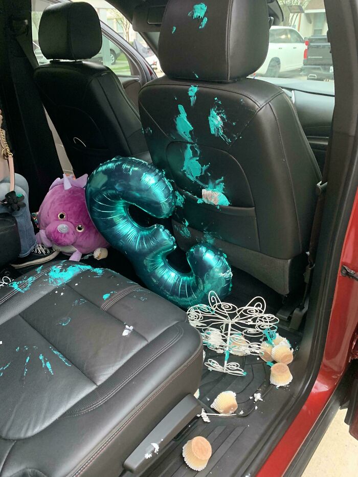 Car interior mess with blue paint splatters, a purple stuffed toy, balloon, and spilled cupcakes on black leather seats.