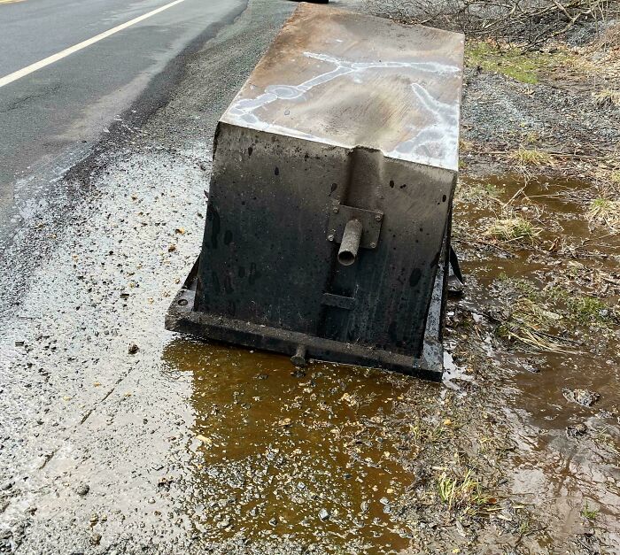 Rusty metal box tipped beside a wet muddy roadside, showing a total mess in an outdoor setting with damp ground.