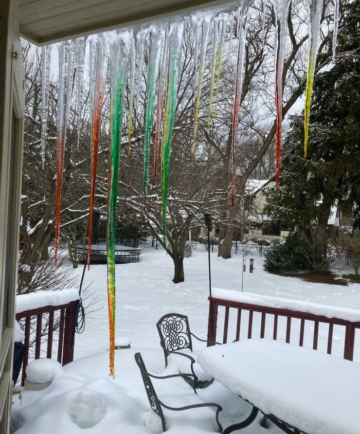 Long colorful icicles hanging from a porch roof over a snowy backyard with outdoor furniture and bare trees.