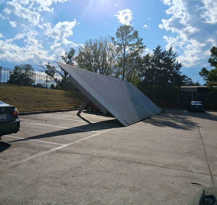 Metal carport collapsed in parking lot, showcasing a total mess and chaos on a sunny day with scattered cars nearby