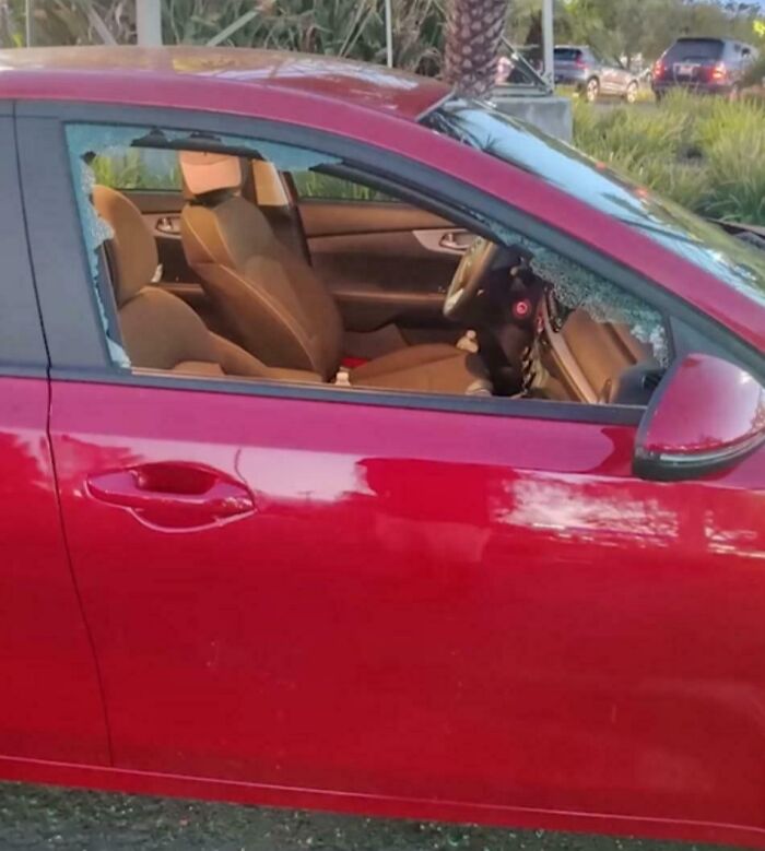 Red car with shattered driver side window parked outdoors on a sunny day, showing interior seats and steering wheel.