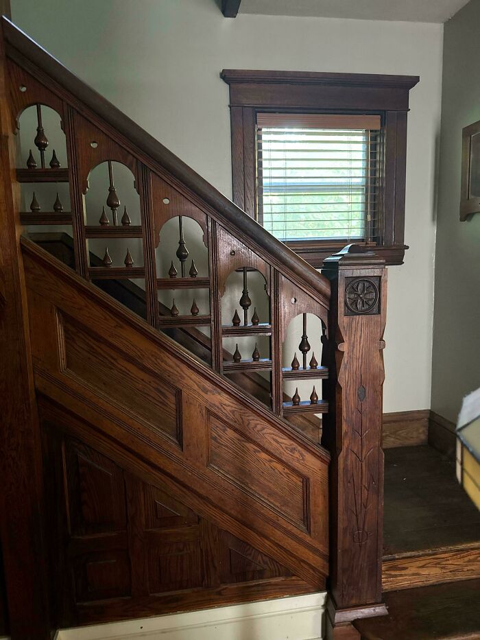 Intricately carved wooden staircase railing in a charming old home with a window and blinds in the background.