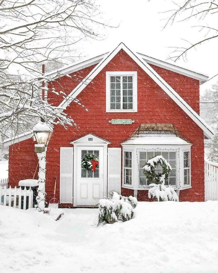 Charming old red cottage covered in snow with wreath decorations on door and window in winter setting.