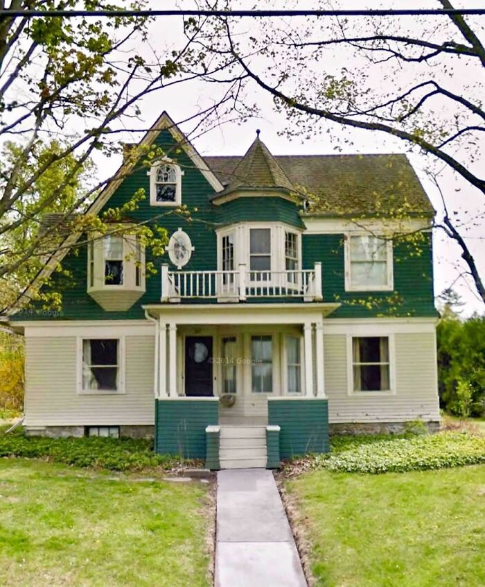 Charming old home with green and white exterior surrounded by spring trees and a walkway leading to the front porch.